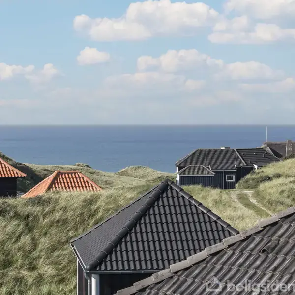 Huse med sorte og røde tage står blandt sandklitter med græs ved havet. Blå himmel med spredte hvide skyer danner baggrunden for den kystnære scene.