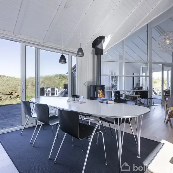 Et moderne spisestuebord med fire stole omkring står på et tæppe. Et ildsted er tændt ved siden af. Store vinduer viser en udendørs terrasse og grønt landskab.