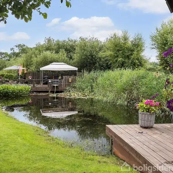 En have med en træterrasse og blomster i en krukke ved siden af en dam. Træer og buske danner en grøn baggrund, med en anden terrasse og parasol i baggrunden.