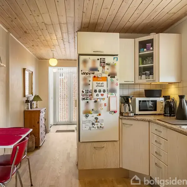 Køkken med lyse trælåger og hvidevarer, herunder køleskab med magneter og mikroovn. Rød bordplads med stol i spisekrog. Træloft og et stort vindue, der slipper lys ind.