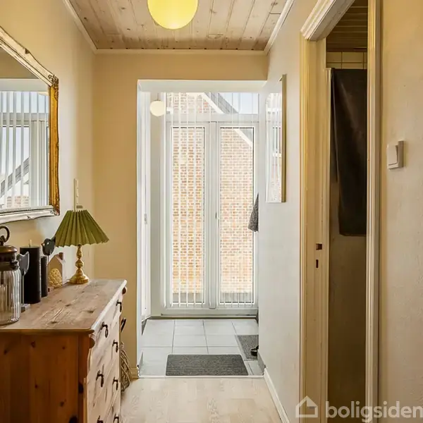 Kommode står i en gang med en lampe og spejl på væggen. Gangen leder til en lys entré med glasdør, der viser en murstensvæg udenfor.