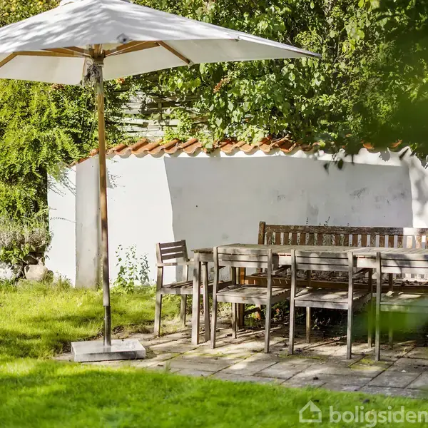 En træterrasse med et sæt havemøbler, herunder borde og stole, står under en stor hvid parasol. Omgivet af grønt græs og en hvid mur med rød kant i sollys.