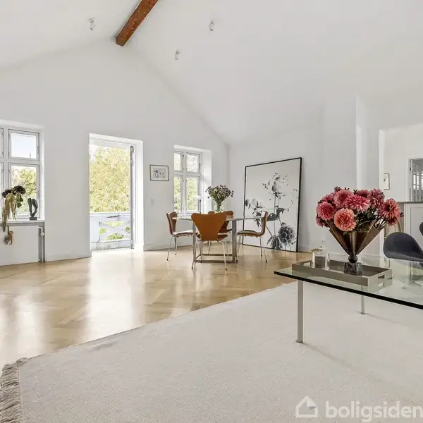 En lys stue med højt loft og træbjælker. Et glasbord med blomster står på et lyst tæppe. Rundt om står moderne møbler, og store vinduer slipper naturligt lys ind.