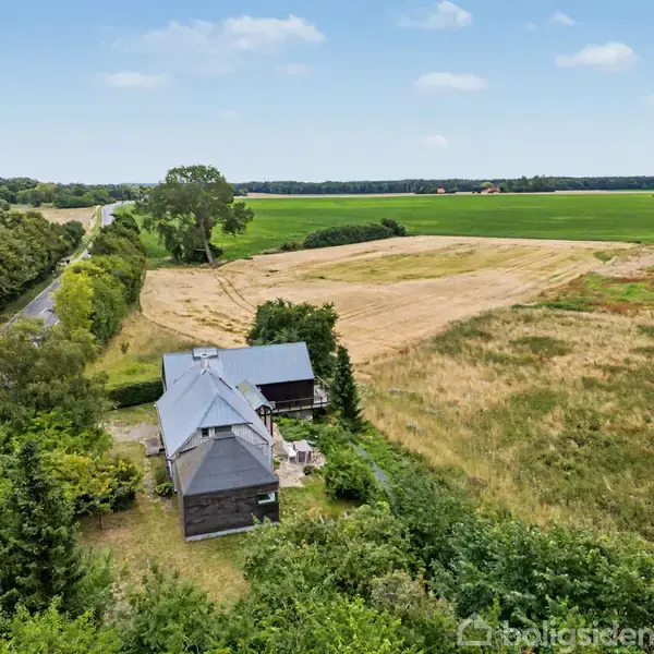 Et hus med gråt tag ligger omgivet af frodig vegetation og åbne marker. En vej løber langs ejendommen med skov i baggrunden, under en let skyet himmel.