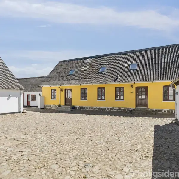 Gult bindingsværkshus med mørkt tag står på en brostensbelagt gårdsplads. Omgivet af hvide bygninger under en blå himmel.