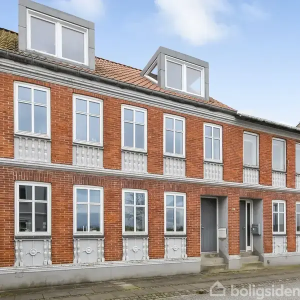 Rødstensbygning med to etager og kviste på taget. Der er hvide vinduer og døre. Bygningen står i en bymæssig gade med fortov og klar himmel i baggrunden.