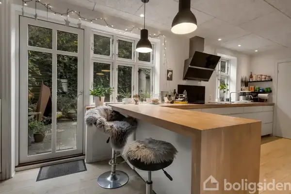 Køkkenbord med barstole dækket af pelstæpper, står i et moderne køkken med hængende lamper. Store vinduer ved siden af døren fører ud til en terrasse med planter.