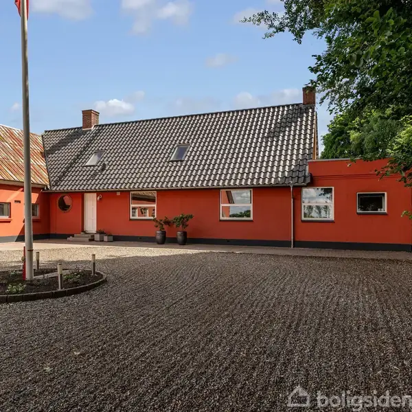 Et rødt hus med klassisk dansk arkitektur står i solen. Foran ses en grusbelagt gårdsplads med en flagstang i midten. Omkring er der træer og blå himmel med skyer.
