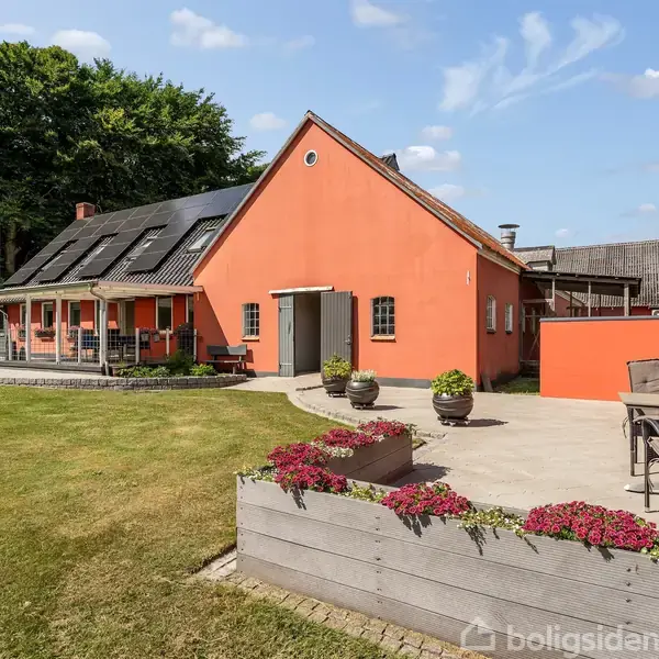 Et orange hus med en sort tagterrasse og en overdækket veranda. Huset står i en velplejet have med græsplæne, flisebelagt terrasse og blomsterkasser fyldt med røde blomster.