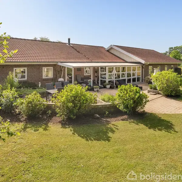 En murstensvilla med rødt tegltag har en overdækket terrasse fyldt med havemøbler. Den er omgivet af en velholdt græsplæne og buske, midt i et frodigt, solrigt haveområde.