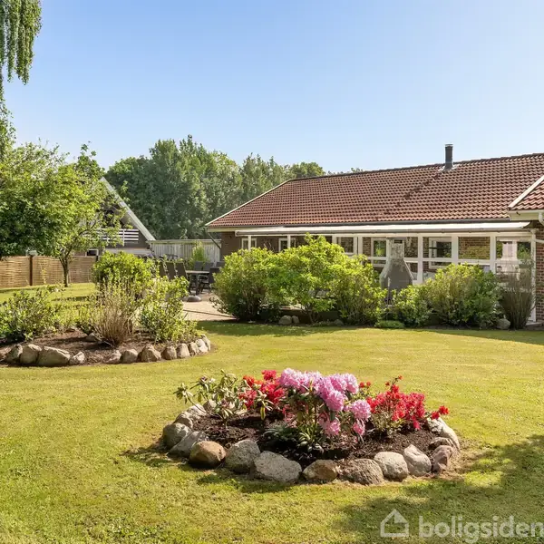 Et murstenshus med rødt tag står i en velplejet have. Haven har grønne plæner, farverige blomsterbede med stenkanter, omgivet af buske og træer under en klar blå himmel.