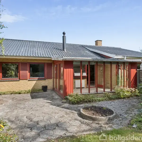 Hus med fladt tag, gule mursten og røde vinduesrammer står på en terrasse omgivet af frodig have. Store vinduespartier vender mod haven, hvilket tillader naturligt lysindfald.
