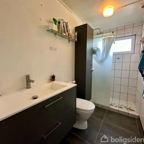 Badeværelse med sort vaskeskab og hvid håndvask. Bruser er i hjørnet med glasdør, flisevægge, og lille vindue. Håndklæde hænger på en krog. Hylde har toiletartikler.