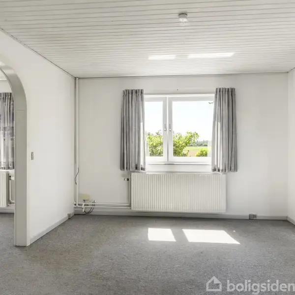 Et tomt rum med lyse vægge har et vindue med gardiner og en radiator nedenunder. Der er en bueformet åbning til et tilstødende rum, og sollys strømmer ind.