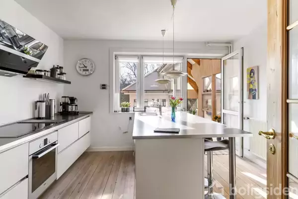 Et moderne køkken har en hvid køkkenø med barstole. En vase med blomster står på øen. Store vinduer og en dør leder ud til en terrasse med udsigt til træer.