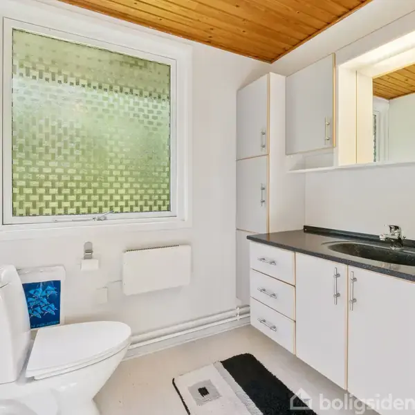 Toilet med hvidt interiør. Vasken er på en sort bordplade med skabe, og et vindue med mønstret glas lukker lys ind. Rummet har lyst træloft og hvide vægge.