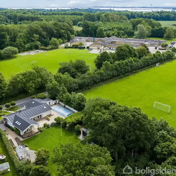 Hus omgivet af skov og store grønne græsarealer, set oppefra. Der er en swimmingpool og en sportsbane med fodboldmål. I baggrunden ses en bygning, muligvis en skole.
