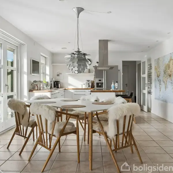 Et spisebord med seks stole dækket af fåreskind står midt i et lyst køkken. En moderne lampe hænger over bordet, med et verdenskort på væggen og store vinduer omkring.