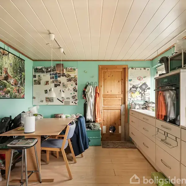 Et køkken med et spisebord og stole til venstre, dekoreret med plakater og billeder på væggene, omgivet af skabe og køkkenudstyr til højre. Møbler og genstande skaber en hyggelig atmosfære.