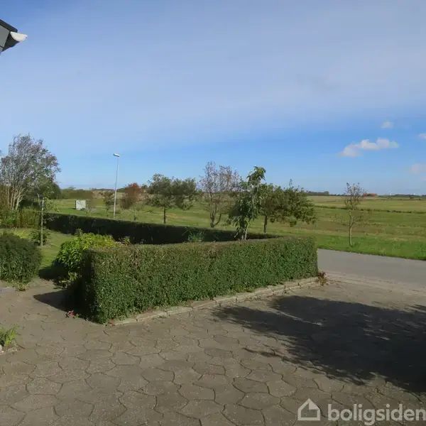 Et hus omkranset af en flisebelagt terrasse med grønne buske og træer, der vokser langs kanten. I baggrunden ses en åben mark under en blå himmel.