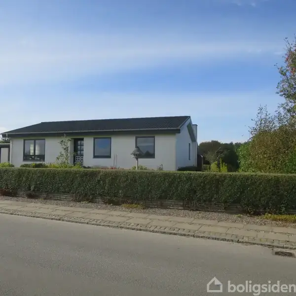 Et hvidt enfamiliehus med sort tag står stille bag en lav hæk. Sollys skinner ned på stedet, og vejen ligger i forgrunden med blå himmel ovenover.
