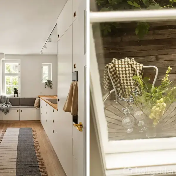 Køkken med hvide skabe, træbordplader og løber, giver et lyst og åbent udseende. Udendørs står et havebord med blomster i vase, set gennem et vindue.