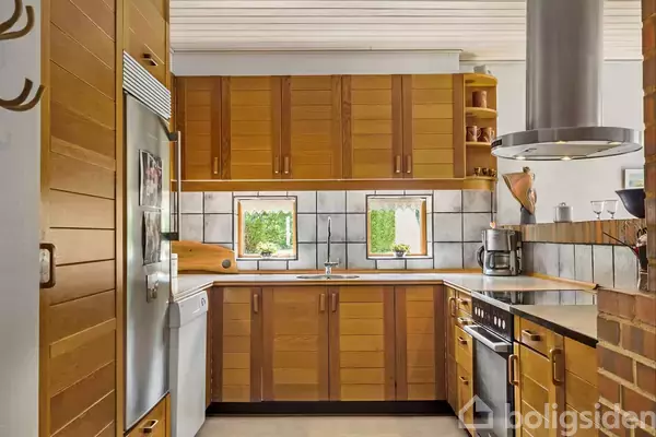 Køkken med træskabe og stålapparater. Et køkkenbord står under to vinduer, der lukker lys ind. Hvid flisevæg omkranser rummet og giver en moderne stemning.