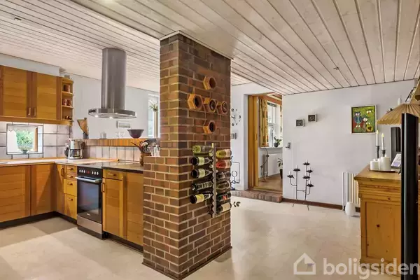 Køkken med træskabe, en ovn med emhætte, og en murstenssøjle med vinopbevaring. Rummet er lyst med hvide lofter og en åben spisestue i baggrunden.