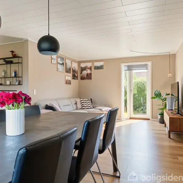 Spisebord med sort bordplade og sorte stole står i en stue med lyse vægge. En vase med røde blomster er på bordet. Stuen har sofa, billeder på væggen, gulvlampe, tv og en stor dør fører ud til haven.