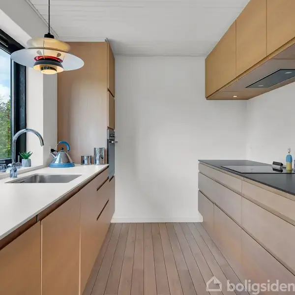 Et moderne køkken med træskabe og en sort bordplade. En vask ved vinduet med udsigt til træer. Loftlampe hænger over arbejdsområdet.