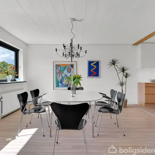 Et rundt spisebord med sorte stole står i en lys spisestue. En moderne lysekrone hænger over bordet. Væggene har farverige malerier. Store vinduer giver udsigt til naturen udenfor.