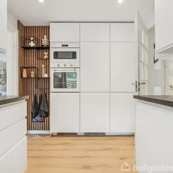 Hvidt køkken med indbygget ovn og mikroovn i højre side, toaster til venstre. Trævæg med hylder og knager til opbevaring. Lyse trægulve og marmorvægdetaljer.