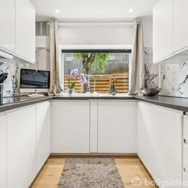 Et moderne køkken med hvide skabe og grå bordplader løber langs væggene. Et vindue med udsigt til en træhegnshave over en vask. En lille tæppe på trægulvet.