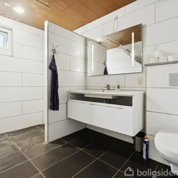 Hvidt badeværelse med væghængt toilet og hvidt skab under vask. Spejl over vasken, hvid flisevæg, sort flisegulv og et lille vindue øverst til venstre.