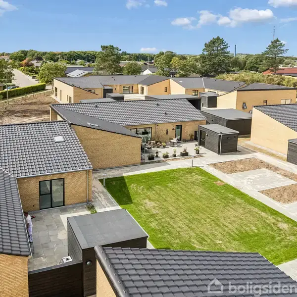 En lys og rummelig stue med sofa og sofabord foran store vinduer. Til venstre er der åbent til spiseplads og køkken, omgivet af moderne, minimalistisk indretning.
