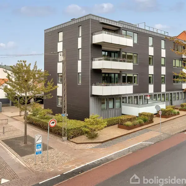 En moderne, sort lejlighedsbygning har flere hvide altaner. Den ligger på et gadehjørne omgivet af fortove og træer. Et cykelstativ og vejskilte er synlige i forgrunden.