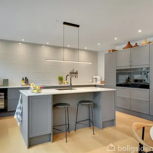 Køkkenø med to barstole og frugtskål står i et moderne køkken. Grå skabe og indbyggede apparater omgiver øen, mens en elegant lampe hænger over. Lyst trægulv danner baggrunden.