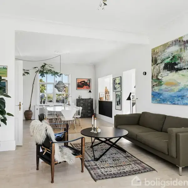 En moderne stue med en grøn sofa og et rundt sofabord. Et tæppe ligger under bordet. Væggene har abstrakte malerier. En stor plante står ved indgangen til spisestuen med udsigt til grønne omgivelser.