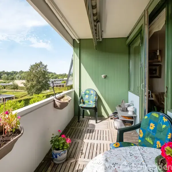 Balkonen er fyldt med farverige stole og et lille bord med blomsterprint. Blomsterkasser pryder rækværket, og omgivelserne består af frodige træer og et blåt himmelstrøg i baggrunden.