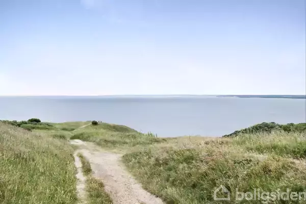 Sti fører ned mod en rolig kyst med udsigt over havet, omgivet af grønne bakker under en klar, blå himmel.
