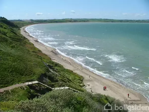 Strandstrækning solbader i solskin, omgivet af grønne skrænter og brusende hav, med enkelte mennesker gående langs vandkanten. En træsti fører ned mod stranden.
