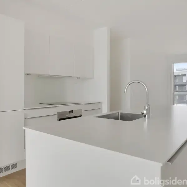 Et moderne, hvidt køkken med en stor køkkenø, indbygget ovn og vask, står i et lyst rum. Gennem vinduerne ses en bygning udenfor.