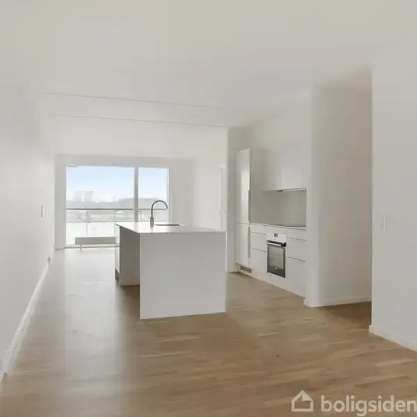 Et moderne køkken-alrum med hvid køkkenø står centralt, med ovn og skabe mod bagvæggen. Rummet har lyse trægulve og store vinduer, der giver udsigt over en by eller landskab.