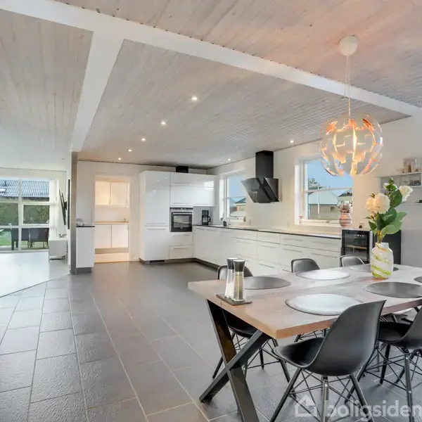 Spisebord med sorte stole står i et moderne køkken-alrum. Køkkenet har hvide skabe og apparater. Store vinduer giver udsigt til en have. En dekorativ lampe hænger over bordet.