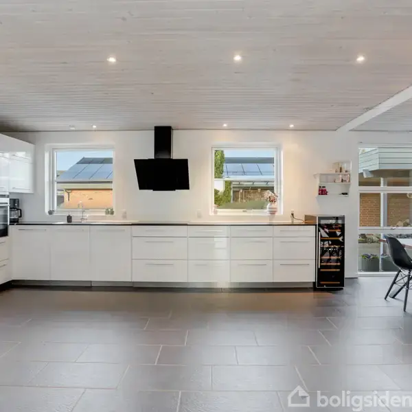 Et moderne køkken med hvide skabe og en sort emhætte, står i et lyst rum med flisegulv. Et spisebord med sorte stole og en dekorativ lampe ses til højre.