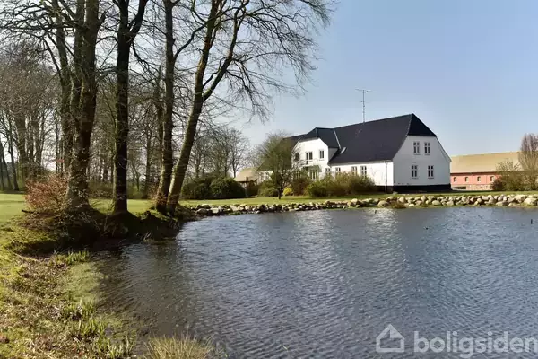 Et hvidt hus står ved siden af en sø omgivet af træer. Landskabet er grønt og landligt, hvilket skaber en fredelig atmosfære.