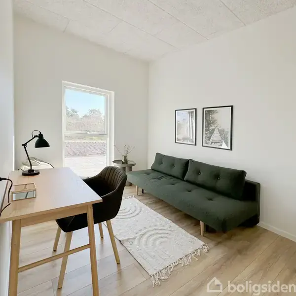 En sofa står i et lyst rum ved siden af et skrivebord med lampe og stol. To billeder hænger over sofaen. Et vindue viser udsigt til en have.
