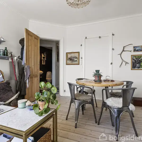 Rundt spisebord med fire stole står i et lyst rum med trægulv. Hylder med bøger og dekorationer findes ved siden af en åben trædør. Indrammede billeder hænger på væggen.