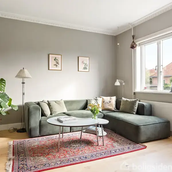 Grå sofa står i en hyggelig, lys stue med trælaminatgulv. Et rundt sofabord står foran den, omgivet af planter og kunst på væggen. Store vinduer slipper naturligt lys ind.