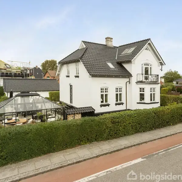 En hvid to-etagers villa med sort tag står ved en vej. Der er en terrasse med glasoverdækning foran huset, omgivet af en velholdt hæk og græsplæne.